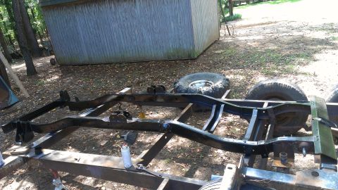 1978 Ford Bronco - The bare frame of a vehicle is set up on stands for work, with tires resting nearby in a wooded area.