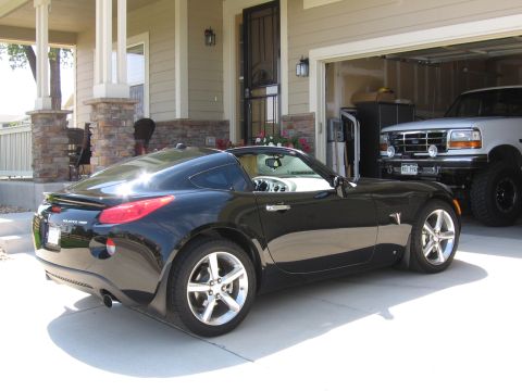 1995 Ford Bronco - A black Pontiac Solstice GXP Coupe is parked in front of a garage, with a lifted Ford Bronco visible inside.