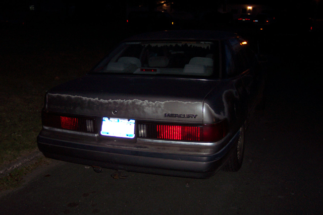 Rear of gray Mercury sedan at night with peeling paint on trunk lid and lit license plate