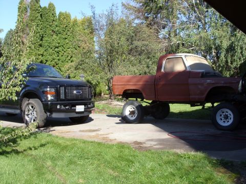 1967 Ford F-250 - A red pickup truck body sits on large wheels next to a black modern pickup truck.