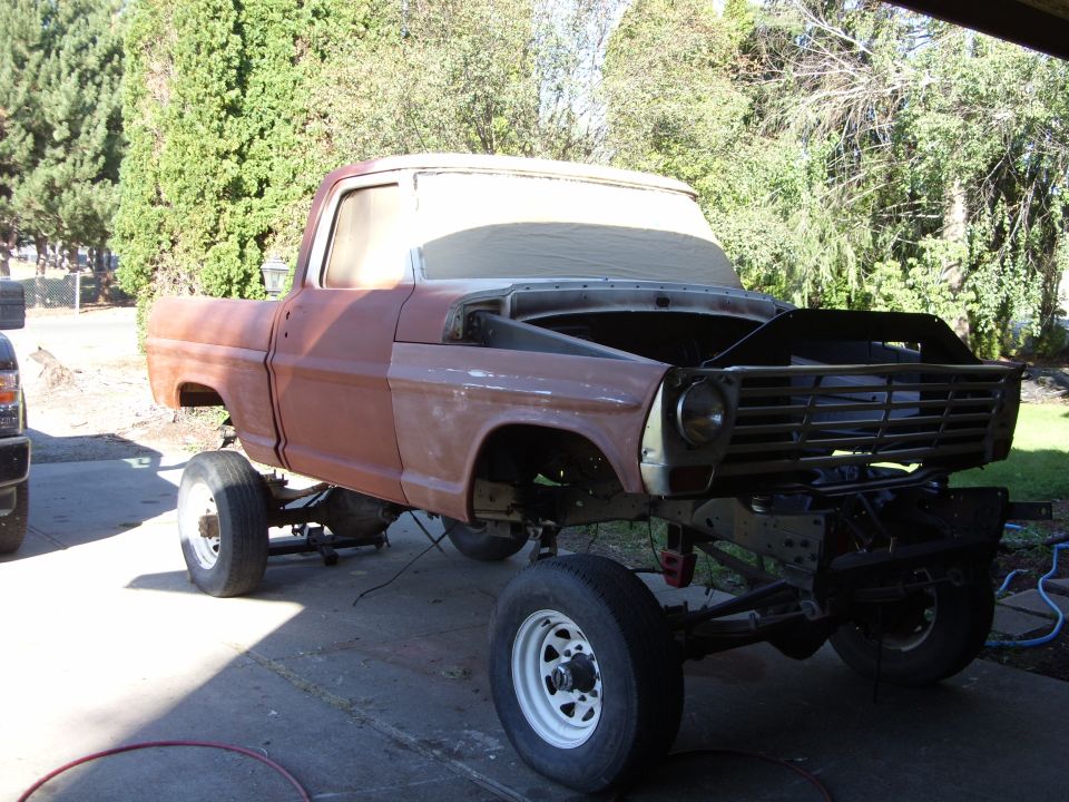 1967 Ford F-250 truck body on jack stands, undergoing restoration. Hood is open, showing engine bay. Front grille is visible