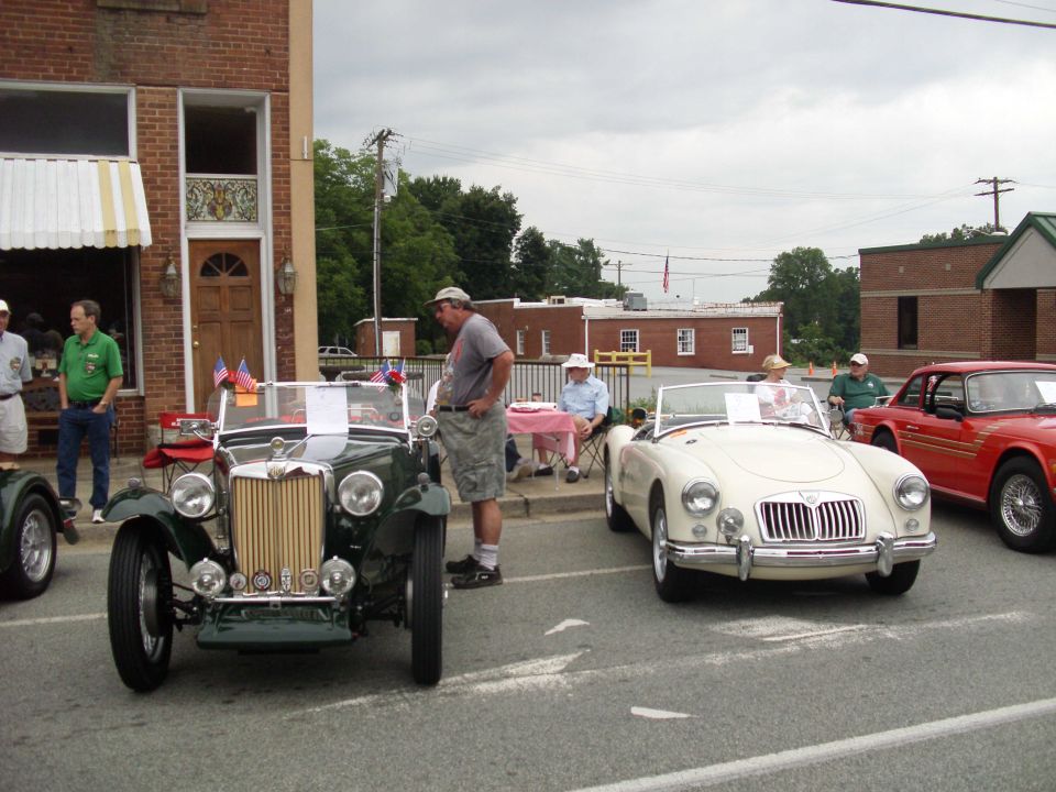 Several vintage convertible cars are parked outside a brick building. People are standing around and sitting at tables