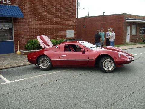 2012 Ford Bronco - A red sports car is parked on the street with its rear hatch open, while a group of men converse nearby.