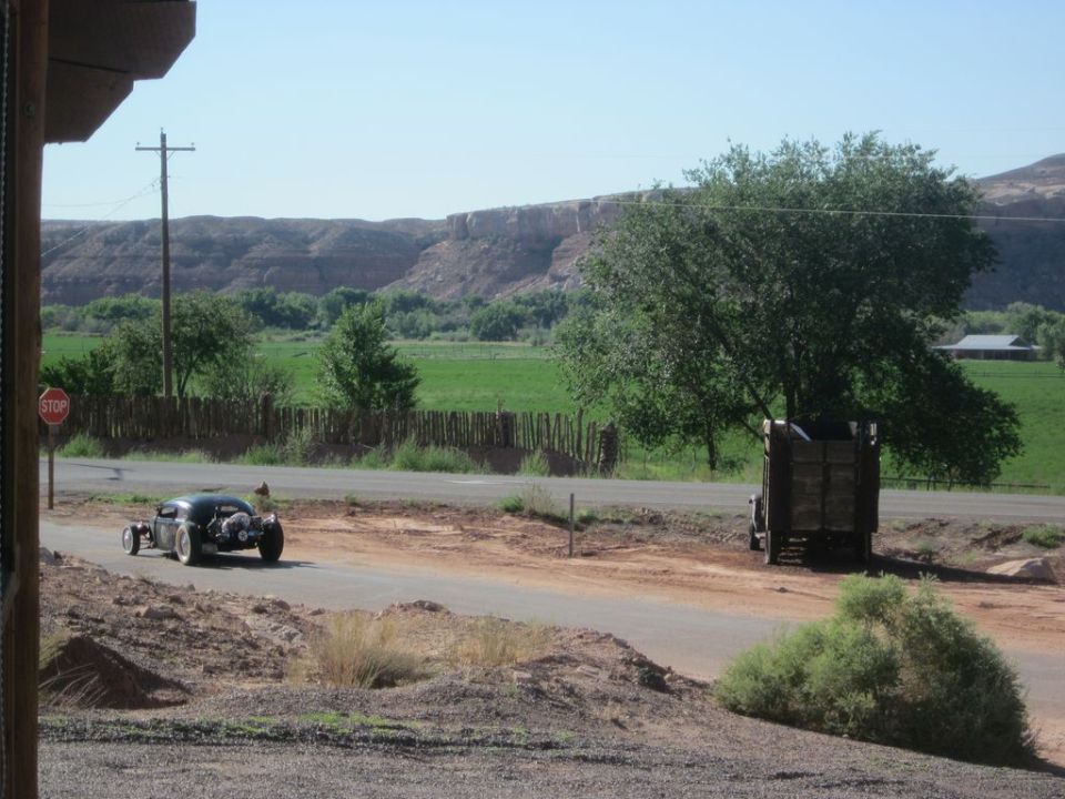 Hot rod car drives past a parked truck on a rural road with a mesa in the background. Supermotors logo visible