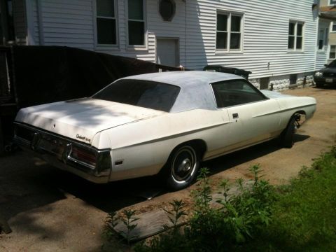 2004 Ford Crown Victoria - This older model Ford sedan, likely from the early 1970s, is parked in a driveway next to a residential building.