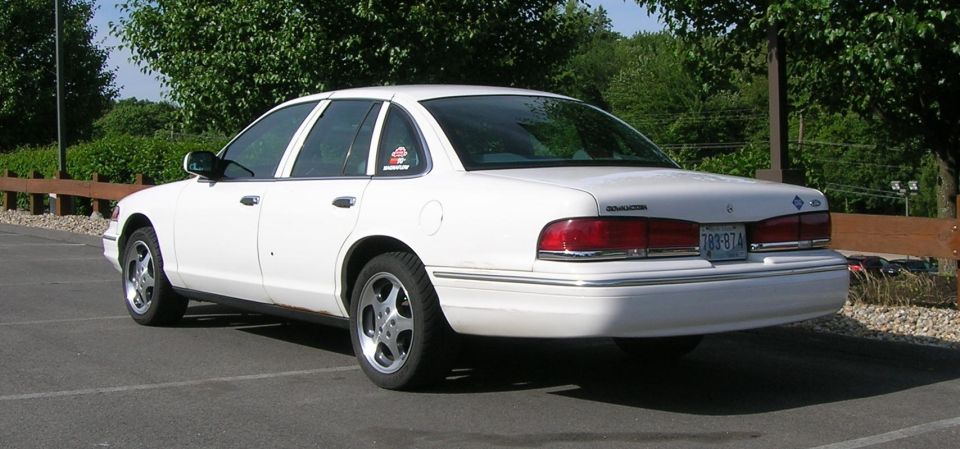 White sedan parked in a lot, rear 3/4 view. Visible chrome trim and aftermarket wheels
