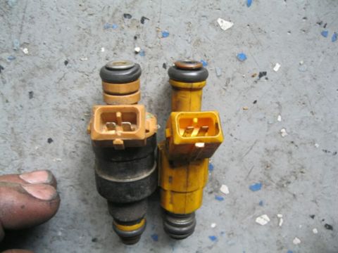 1990 Ford Bronco - Two fuel injectors are shown side-by-side on a concrete surface, with a hand visible on the left.