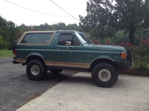 1989 Ford Bronco - This green Ford Bronco, likely a 1987 model, sits on a paved surface with trees and bushes in the background.