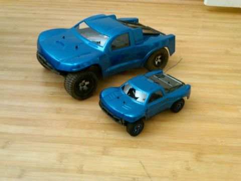 1986 Ford Bronco - Two blue remote-controlled trophy trucks are displayed on a light wood-grain surface. The larger truck is positioned behind and to the left…