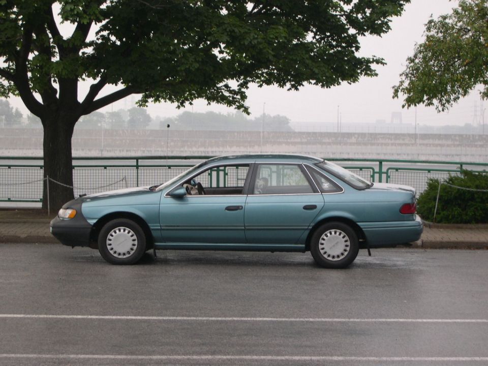 Teal Ford Taurus sedan parked in profile on wet pavement by a tree and waterfront railing