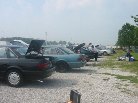 1999 Ford Taurus - Several sedans are parked side-by-side with their hoods and trunks open under a cloudy sky.
