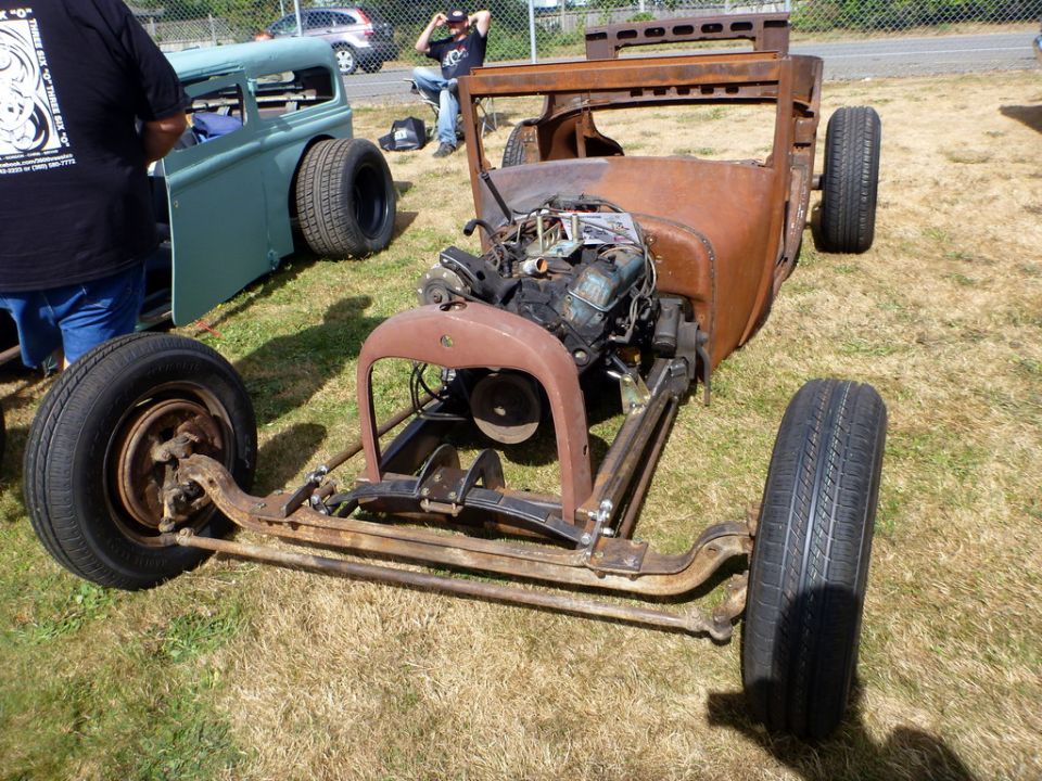Rusty hot rod chassis with engine and front suspension visible. Another car is parked to the left