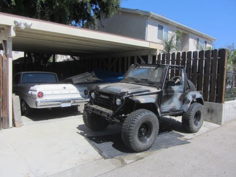 1987 Suzuki Samurai - A Suzuki Samurai with a camouflage paint job and off-road tires is parked in a carport next to another vehicle.