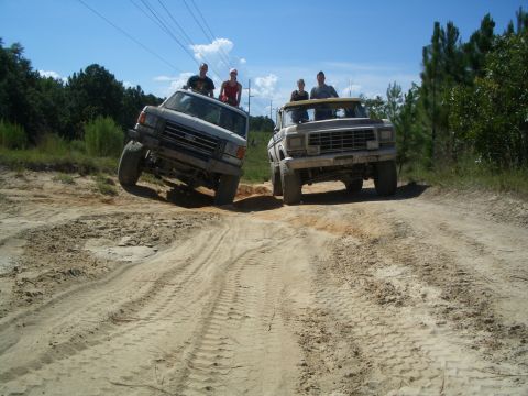 1979 Ford Bronco - Two trucks navigate a sandy trail with passengers riding on top under a clear blue sky.