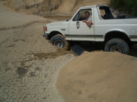 1979 Ford Bronco - A white SUV is stuck in a muddy, cracked terrain, with a large pipe partially buried beneath it.