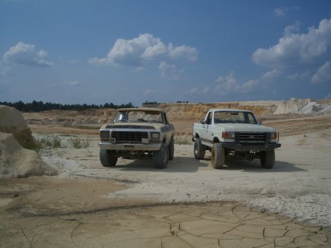 1979 Ford Bronco - Two lifted Ford trucks sit in a dusty, open area with a backdrop of sand hills and trees.