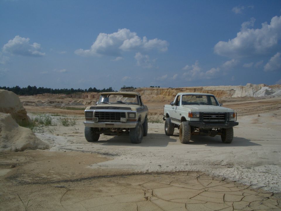 Two lifted Ford trucks parked in a sandy, off-road area under a blue sky with clouds