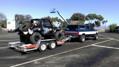 1987 Suzuki Samurai - A modified Suzuki Samurai is transported on a trailer, pulled by a large SUV under a clear blue sky.