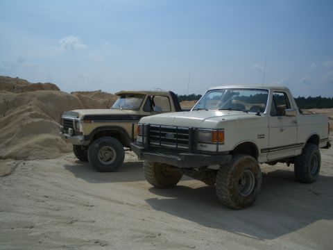 1979 Ford Bronco - Two lifted Ford trucks sit side-by-side in a sandy, outdoor area under a partly cloudy sky.