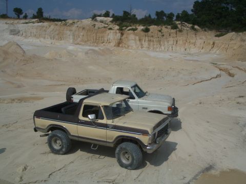 1979 Ford Bronco - A two-tone 1979 Ford Bronco and a white pickup truck sit in a sandy excavation site under a partly cloudy sky.