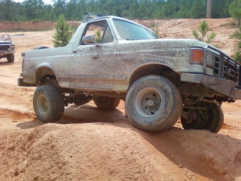 1979 Ford Bronco - A muddy white Ford Bronco navigates a challenging off-road course with a second vehicle visible in the background.