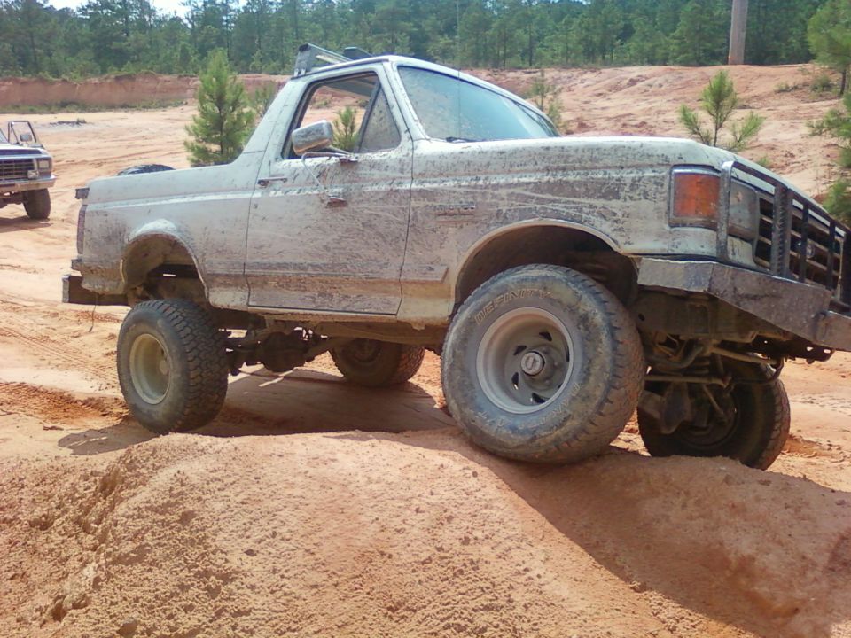 Mud-covered white Ford Bronco on a dirt track, lifted suspension, off-road tires