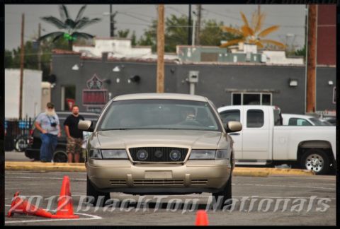 2000 Mercury Grand Marquis - A gold 2000 Mercury Grand Marquis is seen from the front, participating in an event with traffic cones and a white pickup truck in the back…