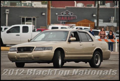 2000 Mercury Grand Marquis - A tan 2000 Mercury Grand Marquis sedan is seen from a 3q front angle, with other vehicles and buildings in the background.