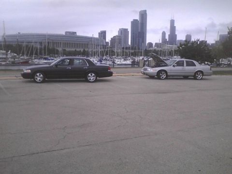 2003 Ford Crown Victoria - Two sedans are parked side-by-side near a harbor with boats and a city skyline.