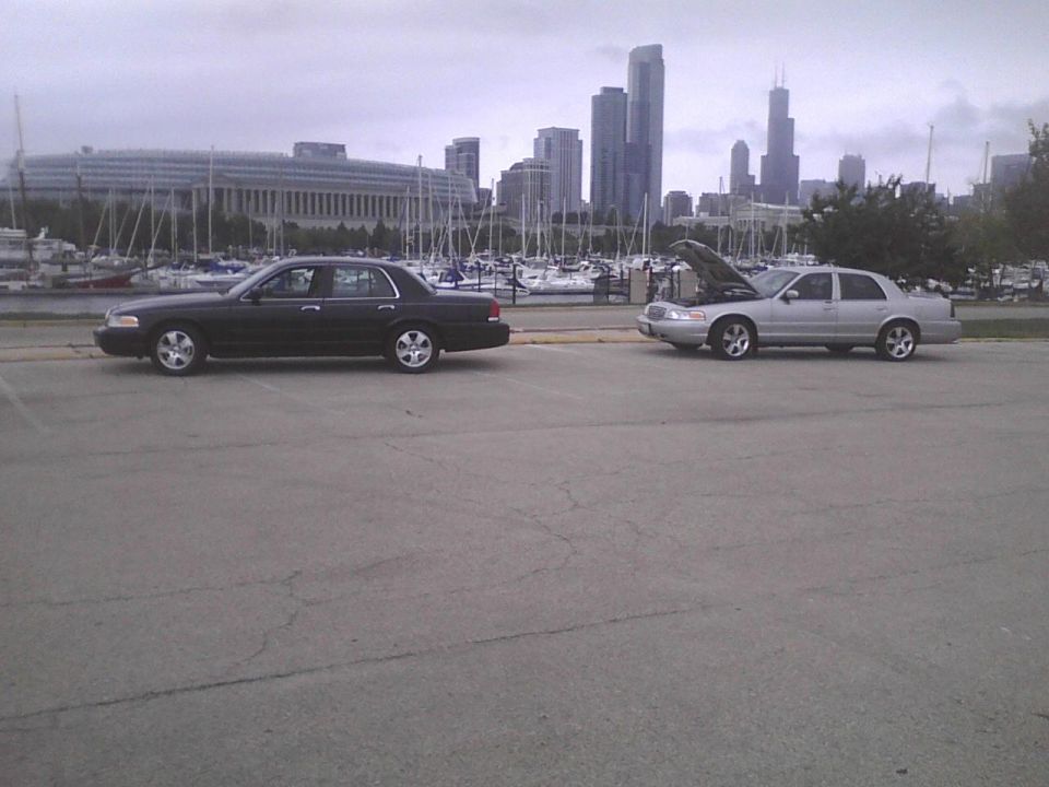 Two sedans parked by a harbor with city skyline in background. One has hood open