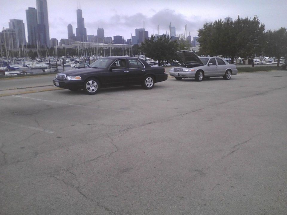 Black 2003 Ford Crown Victoria parked on asphalt, with a silver one behind it with the hood up. City skyline and marina in background