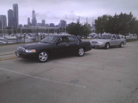 2003 Ford Crown Victoria - Two Ford Crown Victorias are parked in a lot with a marina and city skyline behind them.