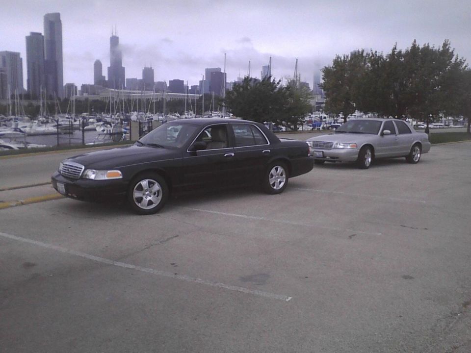 Two black and silver Ford Crown Victorias parked near a marina with a city skyline in the background
