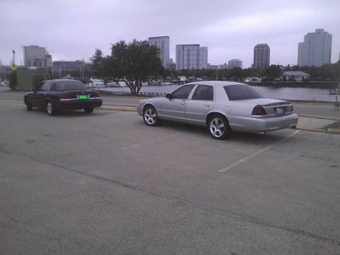 2003 Ford Crown Victoria - Two Ford Crown Victorias are parked in a lot near a body of water with city buildings in the background.