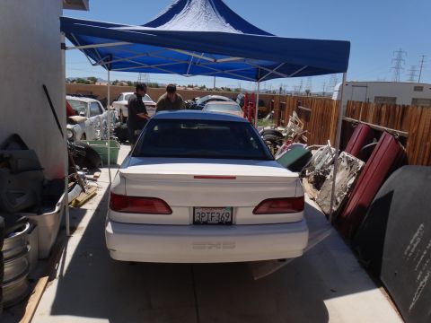 1993 Ford Taurus - A white sedan is parked under a blue canopy in a junkyard with other vehicles and parts visible.
