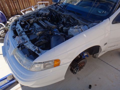 1993 Ford Taurus - A white 1993 Ford Taurus sedan is shown with its hood open, revealing the engine compartment and a removed front wheel.