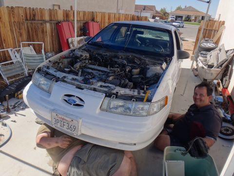 1993 Ford Taurus - Two people are working on a white 1993 Ford Taurus with its hood open in a driveway.