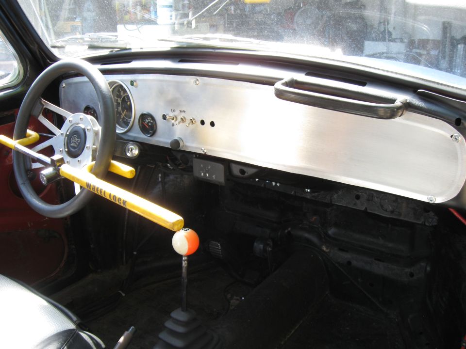 Interior of a VW Beetle showing a custom aluminum dash, aftermarket steering wheel with a yellow wheel lock, and a two-tone shift knob