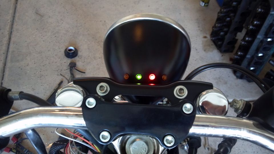 Motorcycle handlebars with a black headlight housing and three indicator lights (two red, one green)