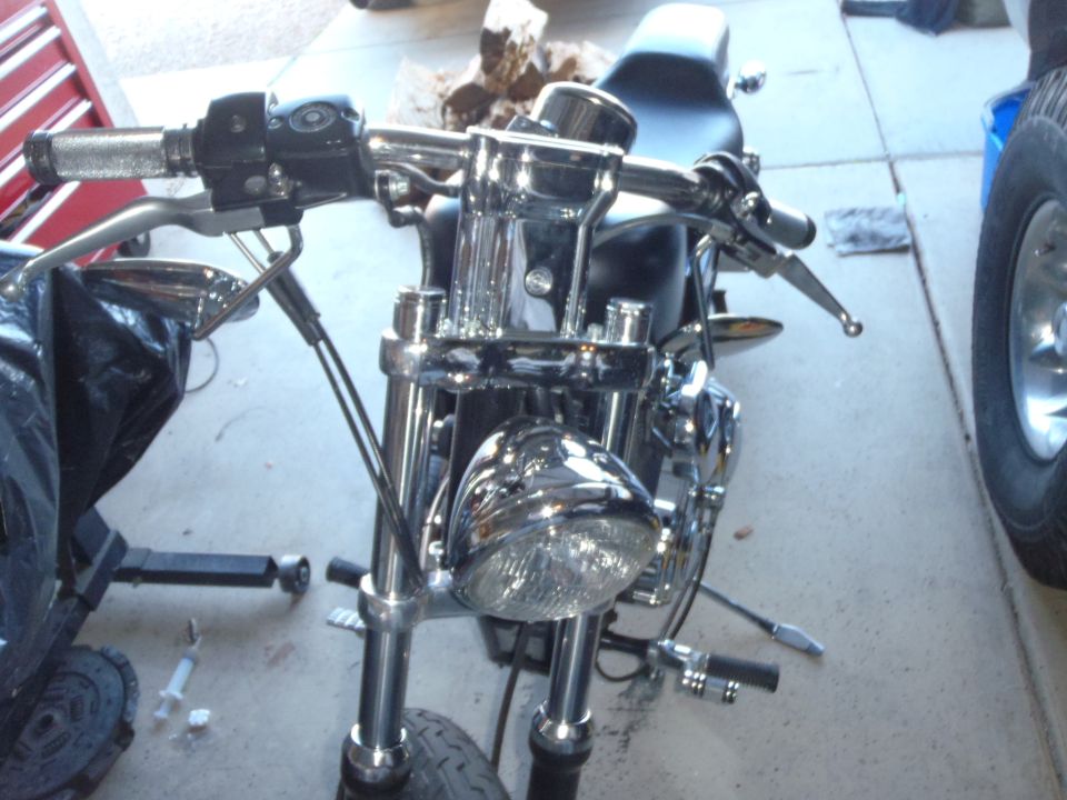 Motorcycle front end with chrome handlebars, headlight, and black seat. A tire is visible on the right