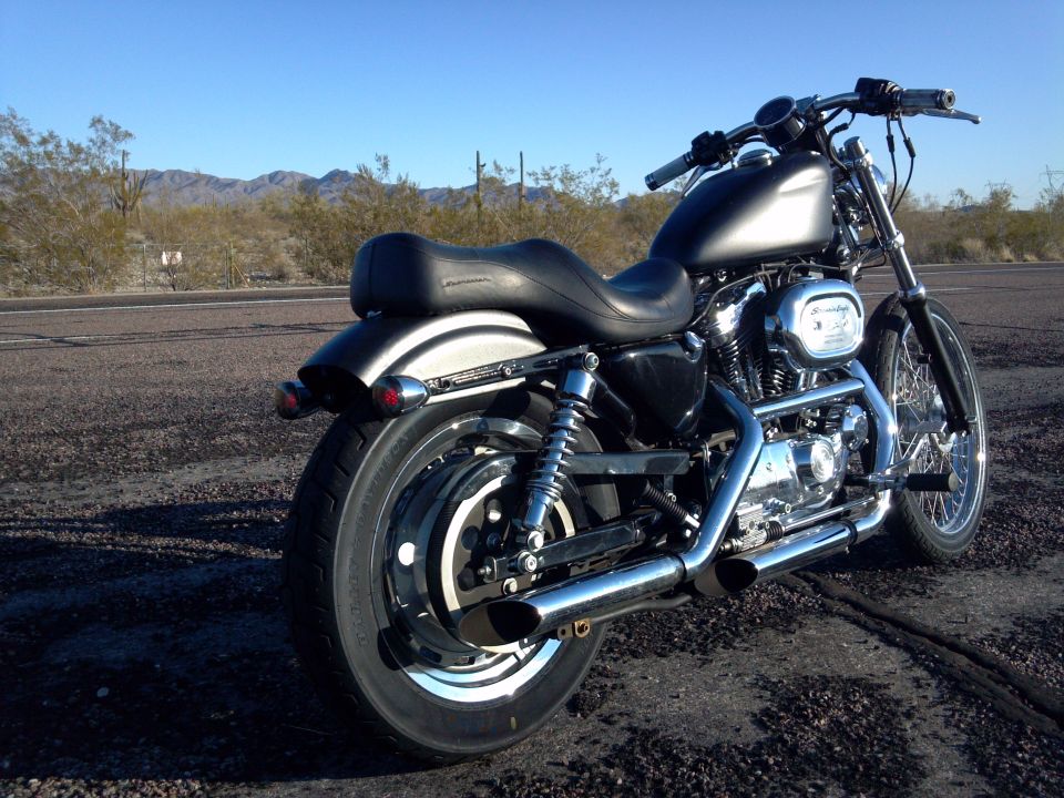 Black Harley-Davidson motorcycle parked on a road with desert landscape behind it. Shows the rear wheel, seat, and engine