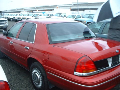 1999 Ford Crown Victoria - A red sedan is parked in a lot with other vehicles under an overcast sky.