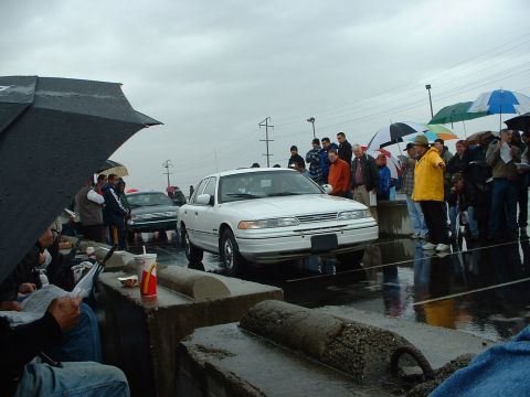 1999 Ford Crown Victoria - A white sedan is presented at an outdoor auction on a cloudy, rainy day with many onlookers.