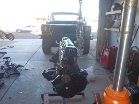 1978 Ford Bronco - An engine is suspended by a chain hoist above a stand, with a truck in the background.