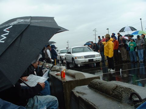 1999 Ford Crown Victoria - A white 1999 Ford Crown Victoria is seen at an outdoor auction event on a rainy day, with people holding umbrellas.