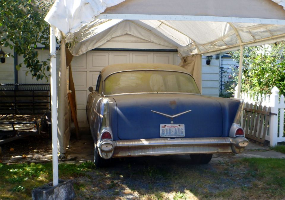 Blue 1957 sedan parked under a carport, rear view. Dusty, with chrome bumper and tail fins