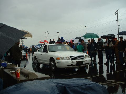 1999 Ford Crown Victoria - A white sedan is displayed at an outdoor auction on a rainy day, with many people holding umbrellas.