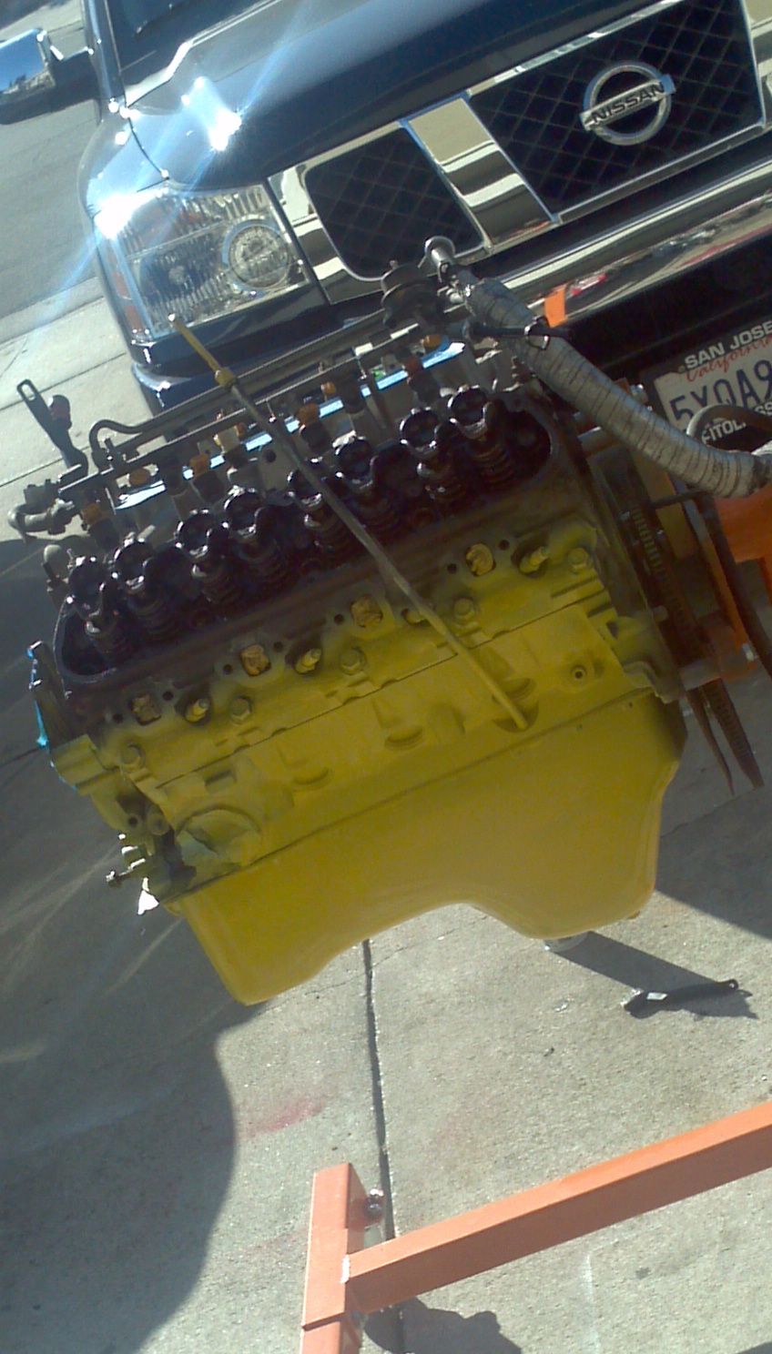 Yellow engine on an engine stand, with the front of a dark Nissan Titan truck behind it. License plate visible