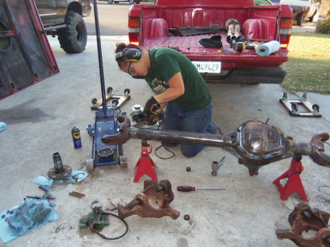 1968 Ford Bronco - A person works on a differential axle assembly in a driveway, with a red pickup truck nearby.