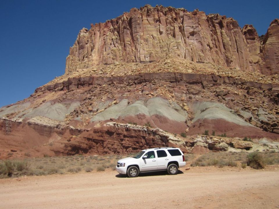 White SUV parked on dirt road with large, layered rock formation in background
